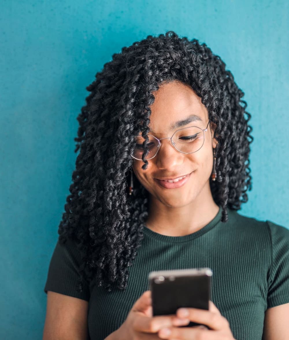 A woman wearing glasses smiling warmly while using her smartphone